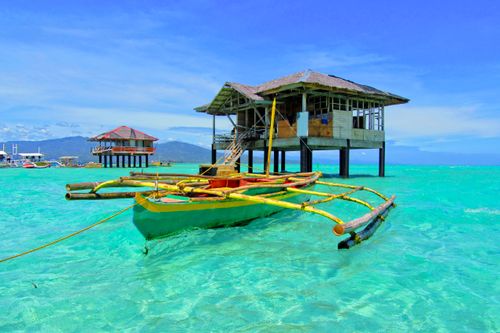 Free A vibrant tropical scene featuring a traditional boat and stilt house over clear turquoise waters. Stock Photo