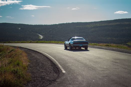 Retro automobile driving down asphalt road next to slopes covered with lush dark green woodland under light blue sky in sunlight