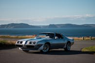 在淡蓝色的天空下，一辆古老的豪华复古跑车停靠在靠近大湖和山脉海岸线的平坦柏油路上