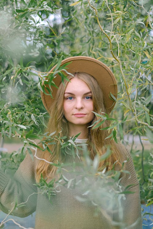 Young attractive female in sweater and hat standing amidst lush green tree branches in garden and looking at camera