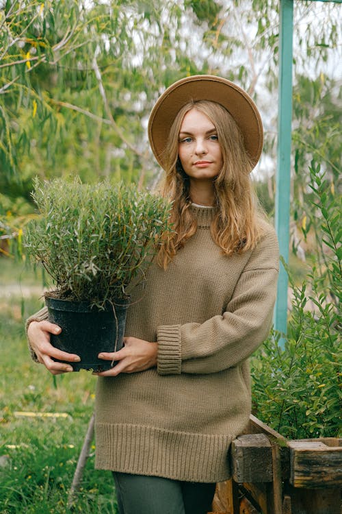 Young positive female gardener wearing brown sweater and hat holding lush potted plant while standing in verdant botanical garden and looking at camera