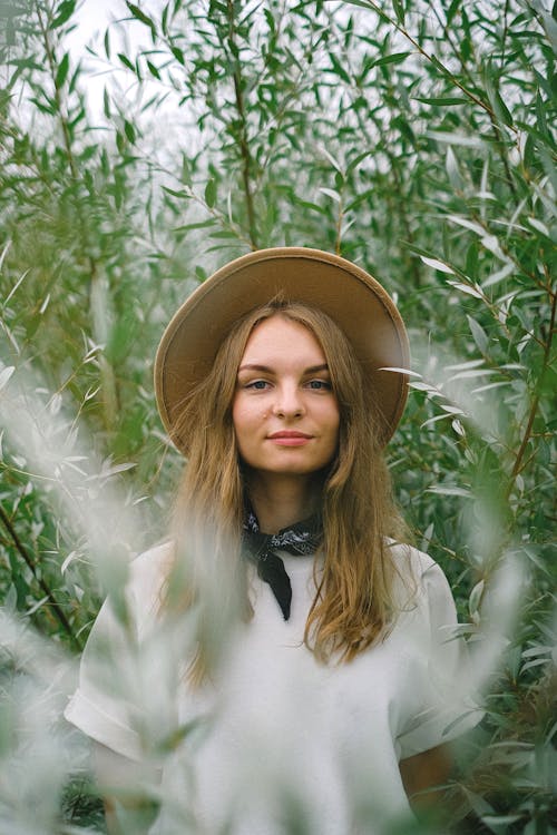 Female farmer wearing hat standing among tall plants in lush farm and looking at camera