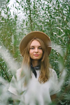 一位戴着帽子的女农民站在郁郁葱葱的农场里，在高大的植物之间，望着镜头