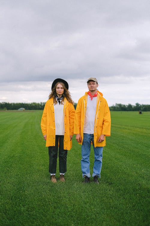 Full body of couple in raincoats standing together in green field with grass under cloudy sky in countryside