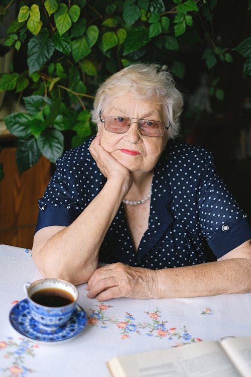 Calm elderly female in casual outfit and eyeglasses looking at camera and leaning on hand while sitting at table with book and cup of tea