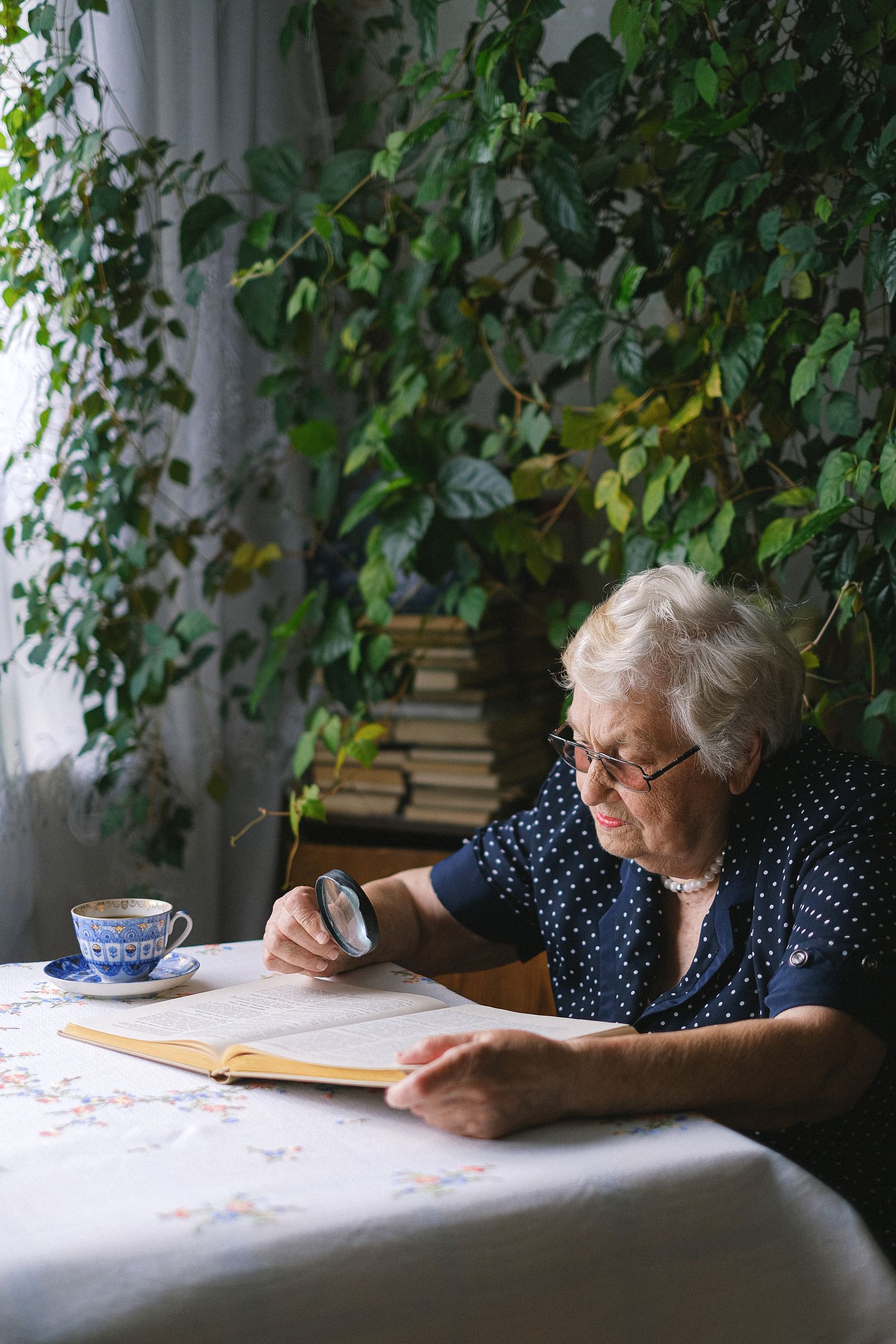 An older woman writing at a table by a window full of houseplants, a cup of tea beside her