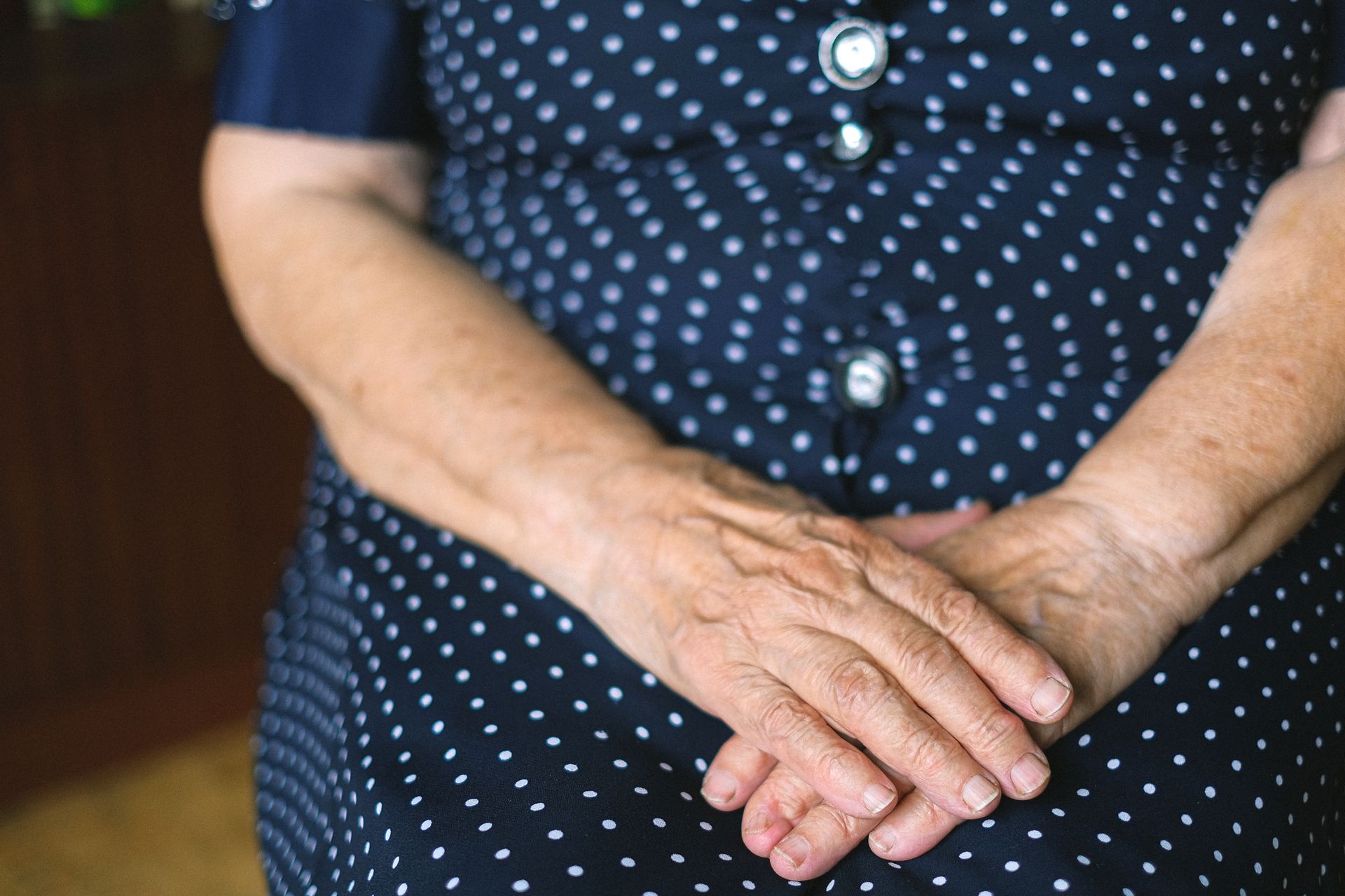 Unrecognizable old female wearing casual polka dot dress with folded wrinkled hands sitting at home in room on blurred background