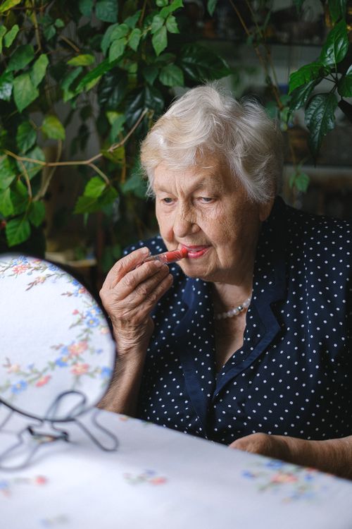 Free Concentrated aged woman with gray hair putting makeup and looking at mirror while sitting at table in room near green plants Stock Photo
