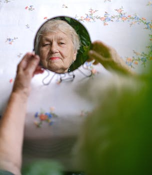 From above of aged female with red lips looking at reflection in mirror while sitting at table with tablecloth in room