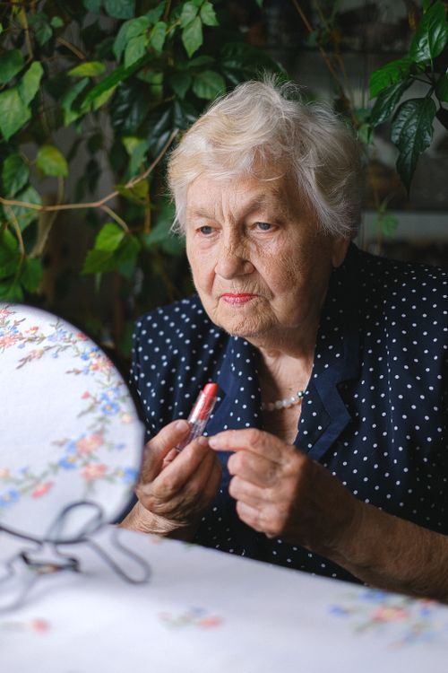 Free Focused aged female with gray hair applying red lipstick and looking at camera while sitting at table near green plants Stock Photo