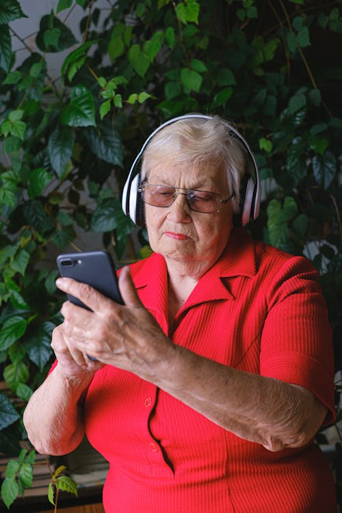 Free Focused aged female wearing red outfit in eyeglasses and headphones listening to music while browsing cellphone in room with green plants Stock Photo