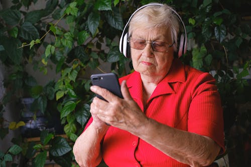 Elderly woman in headphones using smartphone near green leaves
