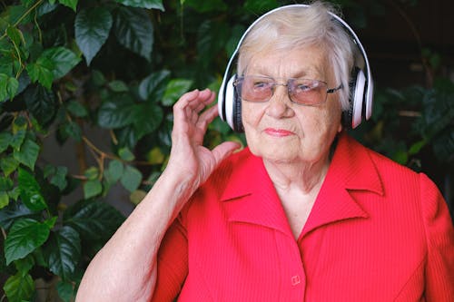 Free Senior thoughtful female in eyeglasses and wireless headphones listening to music near green leaves Stock Photo