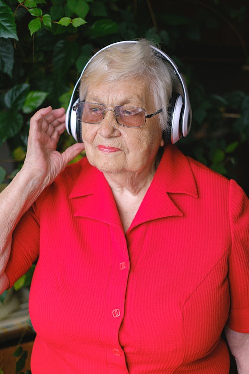 Free Elderly dreamy female touching wireless headphones while listening to music and looking away in garden Stock Photo