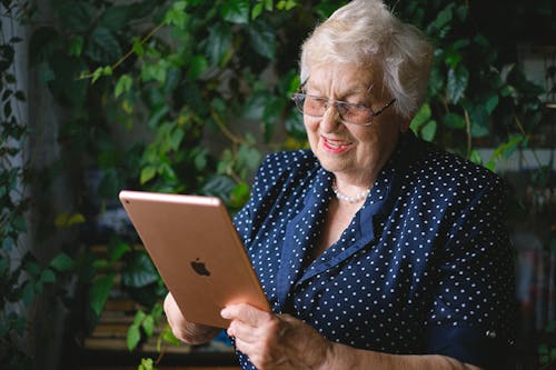 Aged cheerful female smiling and using contemporary tablet while talking on tablet in living room with green plant