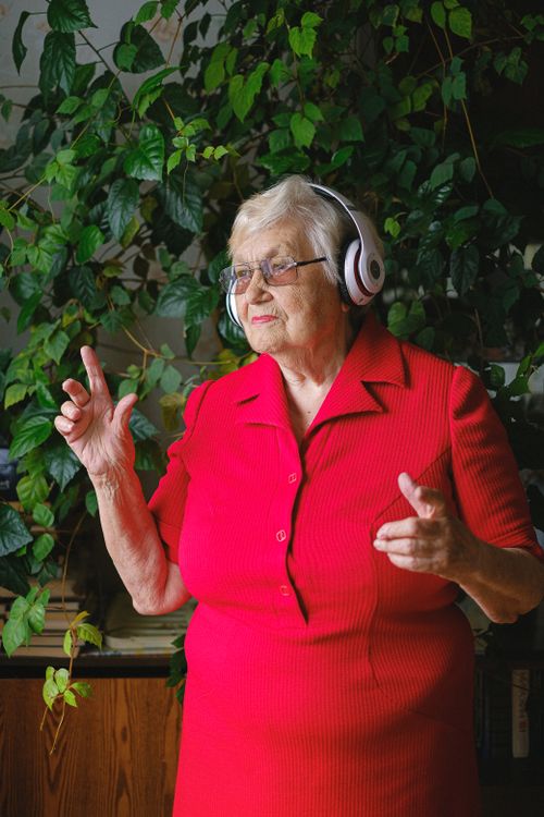 Free Elderly cheerful female in bright red dress and headphones listening to music while dancing and looking away near green plant Stock Photo