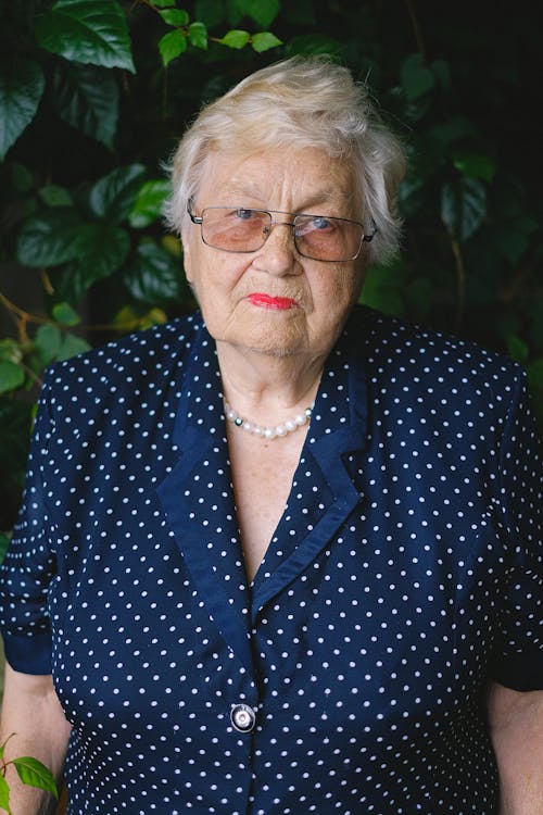 Elderly female with red lips in eyeglasses and polka dot dress looking at camera near plants with green leaves