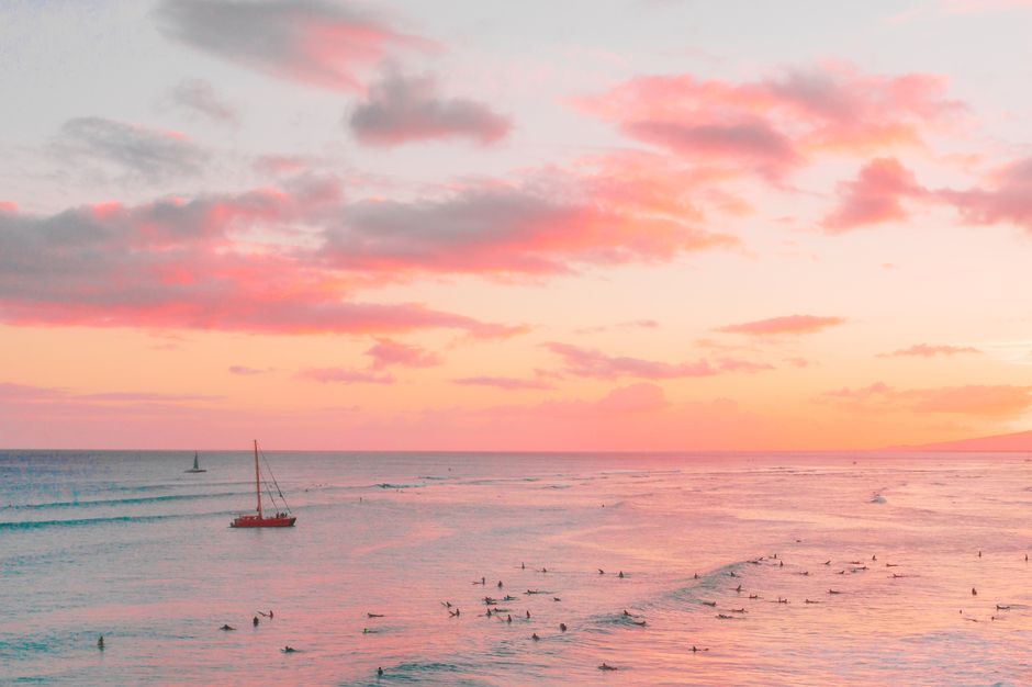 A stunning aerial view of surfers riding waves under a vibrant sunset sky, with boats in the distance.