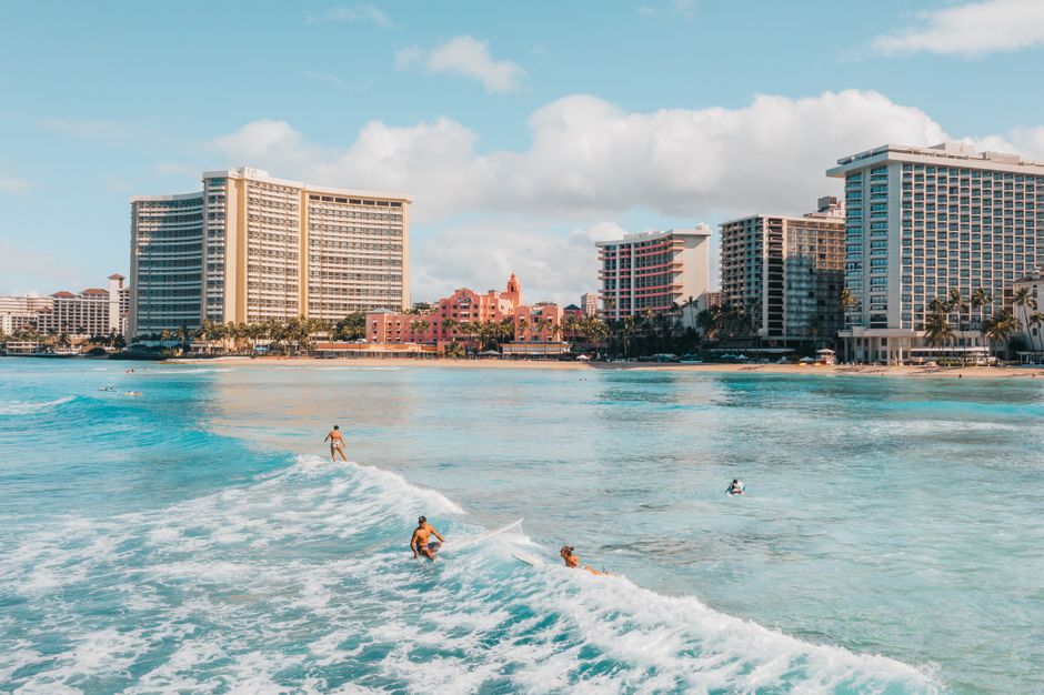 Photo by Jess Loiterton Surfers catch waves with iconic Waikiki hotels in the background, showcasing tropical paradise vibes.
