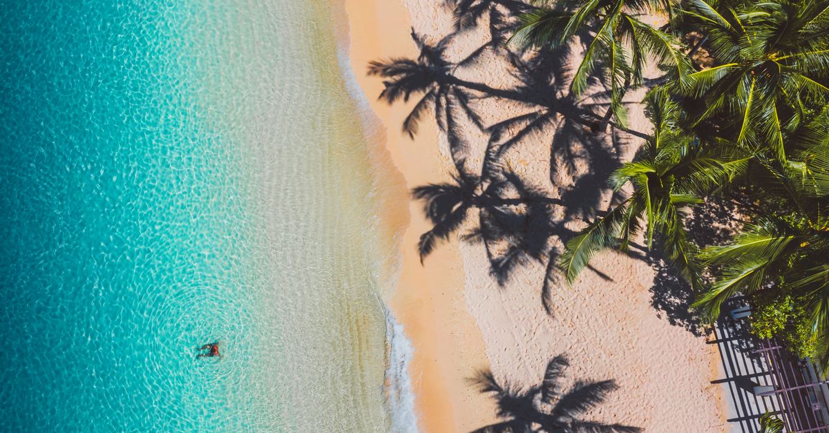 A stunning aerial view of a tropical beach with clear turquoise waters and palm tree shadows on the sand.