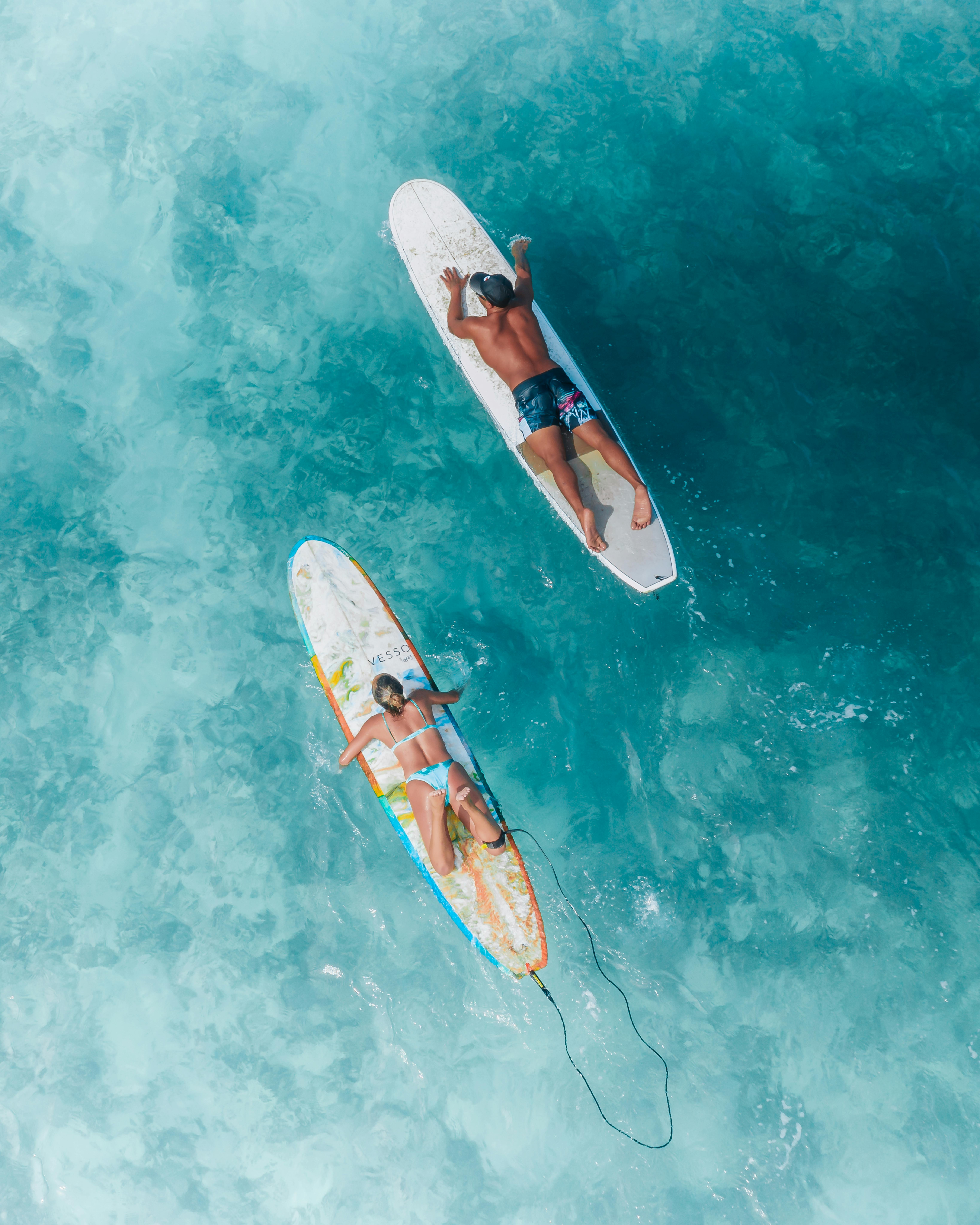 Aerial shot of two surfers relaxing on boards in crystal clear blue ocean water during summer.