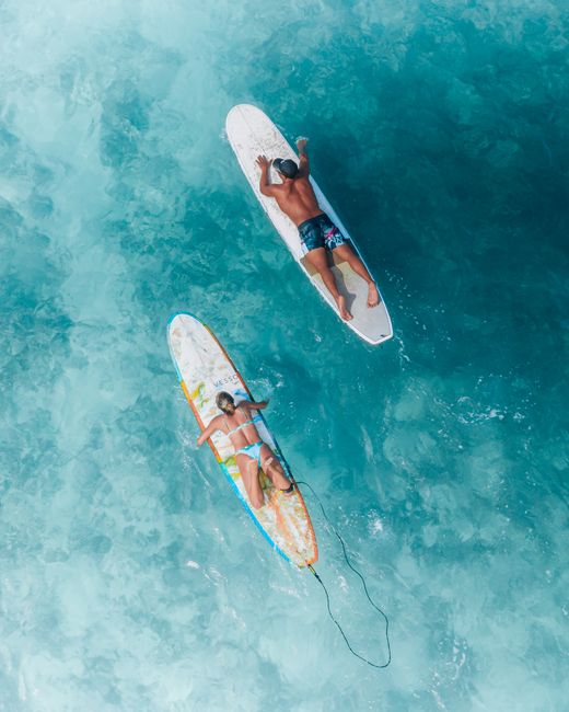 Aerial shot of two surfers relaxing on boards in crystal clear blue ocean water during summer.