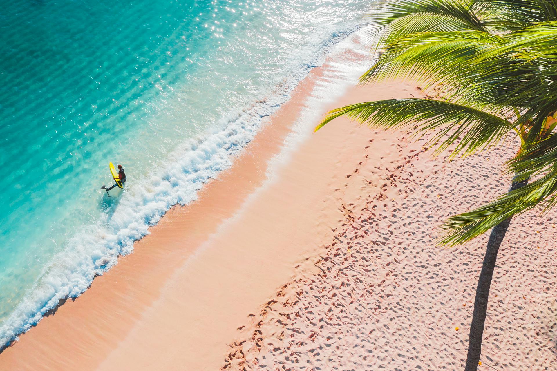 High-angle view of a lone surfer with a board on a tropical beach with turquoise waves.