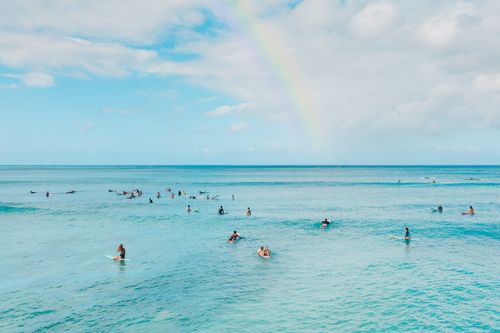 Free Surfers ride gentle waves under a bright sky with a rainbow, creating a lively coastal scene. Stock Photo