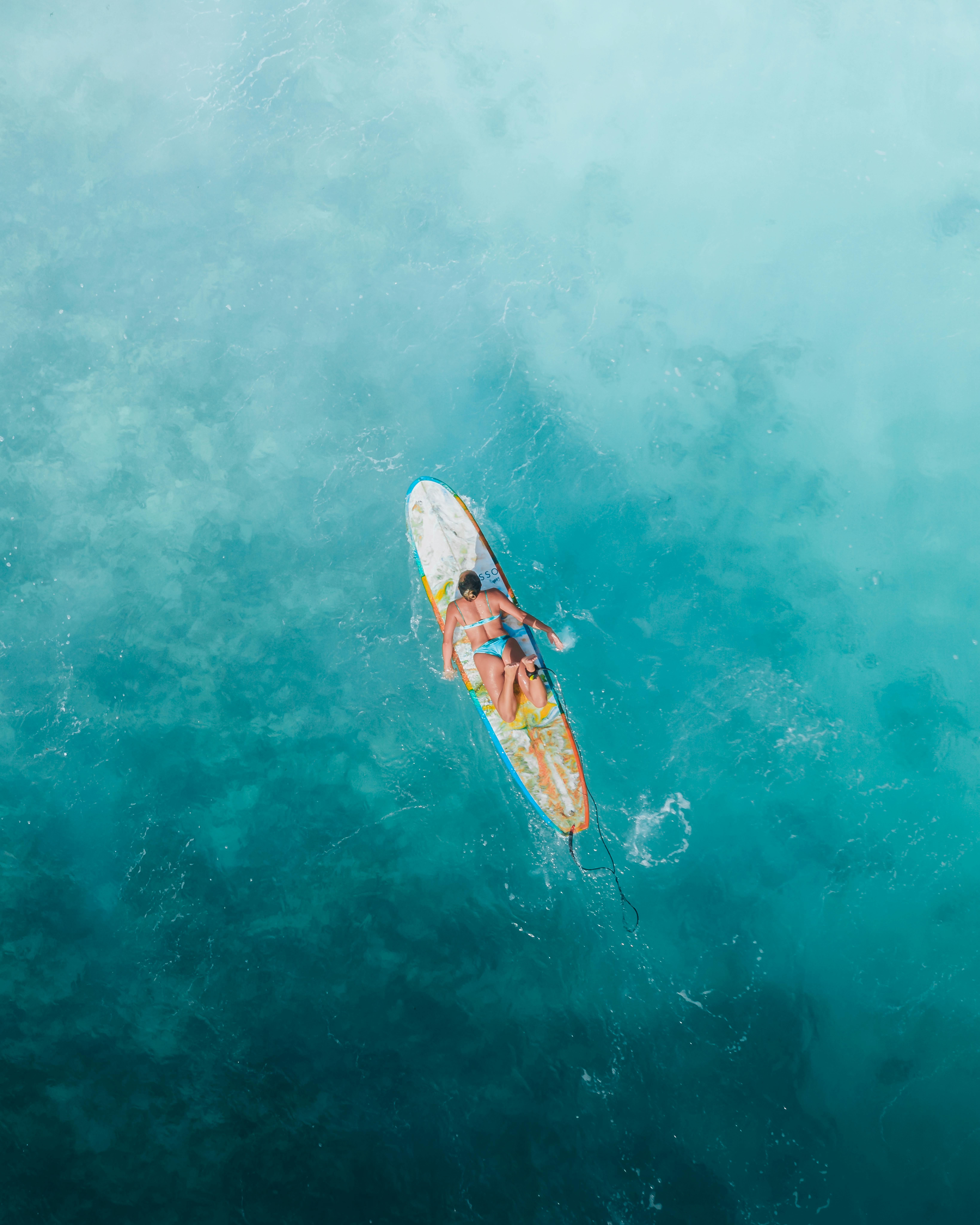 Free Aerial shot of a woman in a bikini relaxing on a surfboard in clear blue ocean waters, ideal for summer vacation themes. Stock Photo