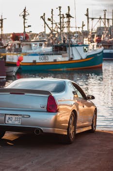 日落时分，一辆豪华跑车停在宁静的港口旁，港口边停泊着渔船