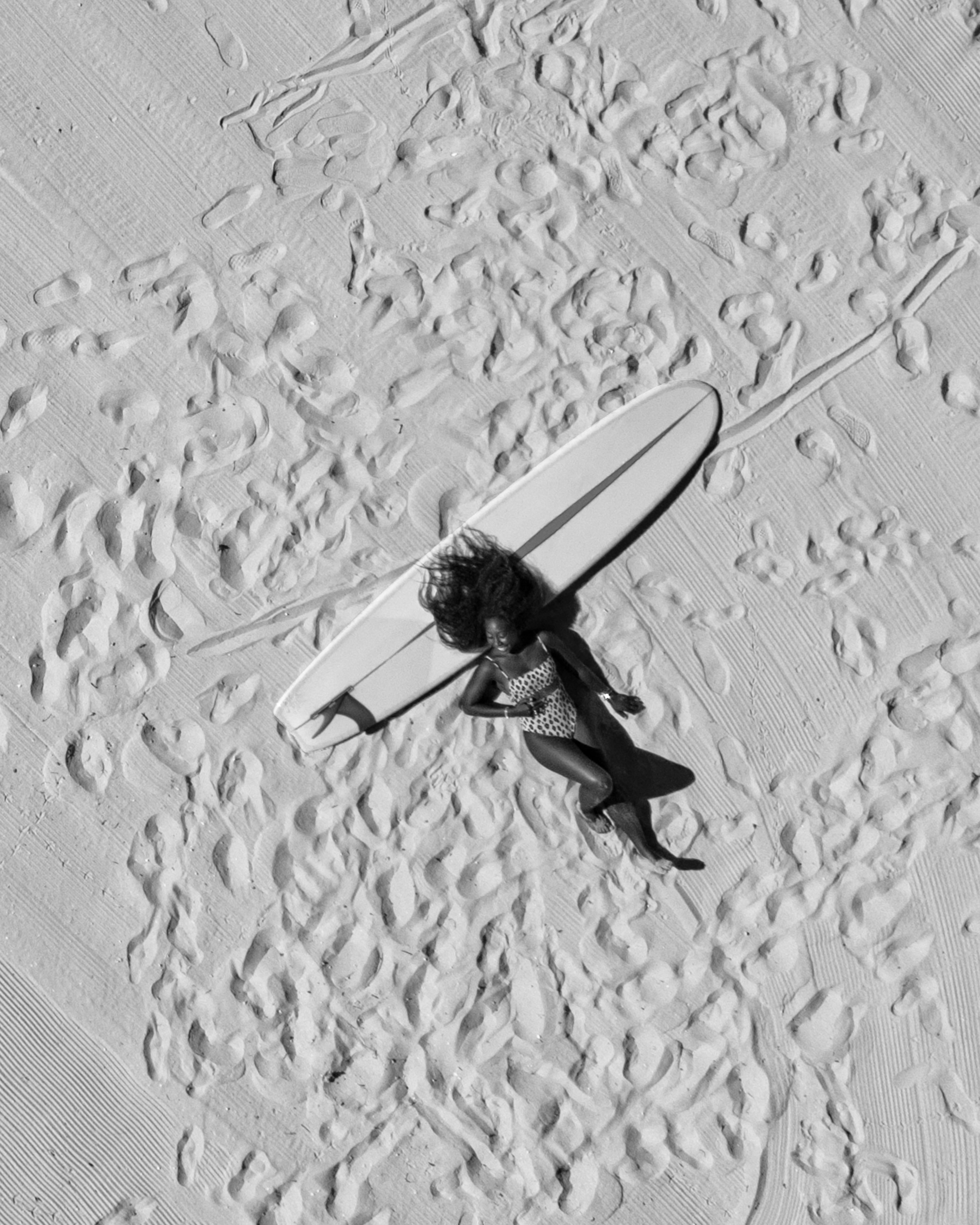 Black and white aerial image of a woman and surfboard on a beach, surrounded by footprints.