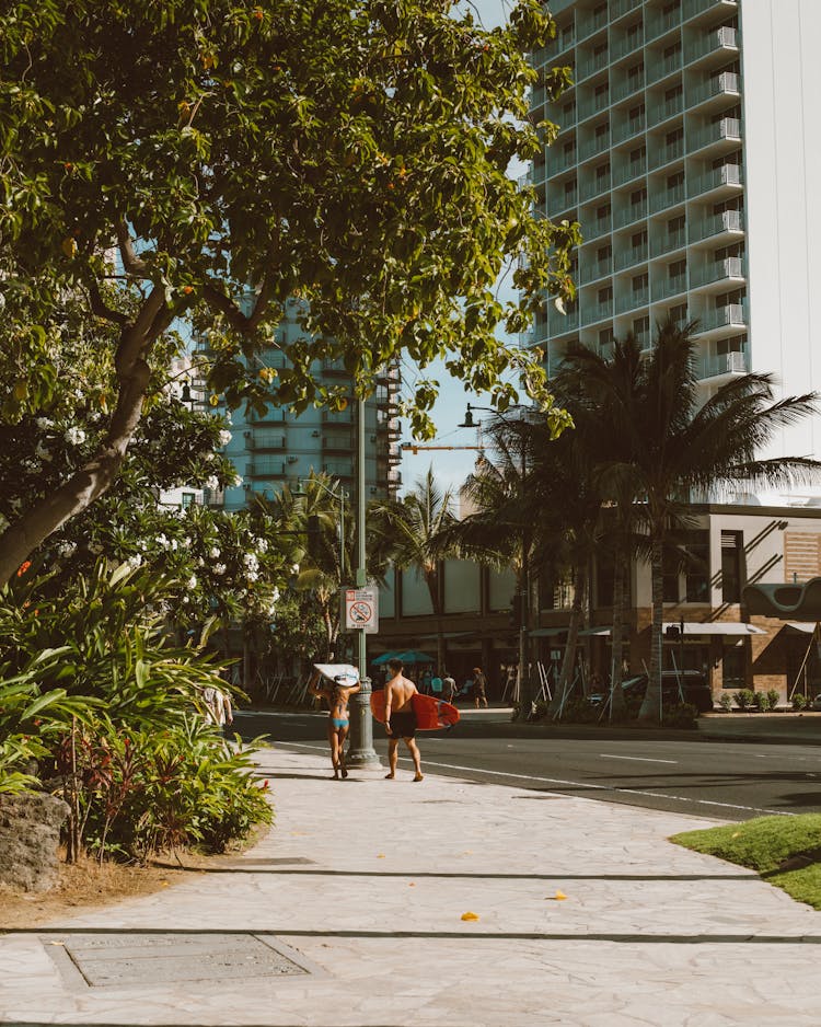 A Man And A Woman Carrying Surfboards While Walking On The Sidewalk