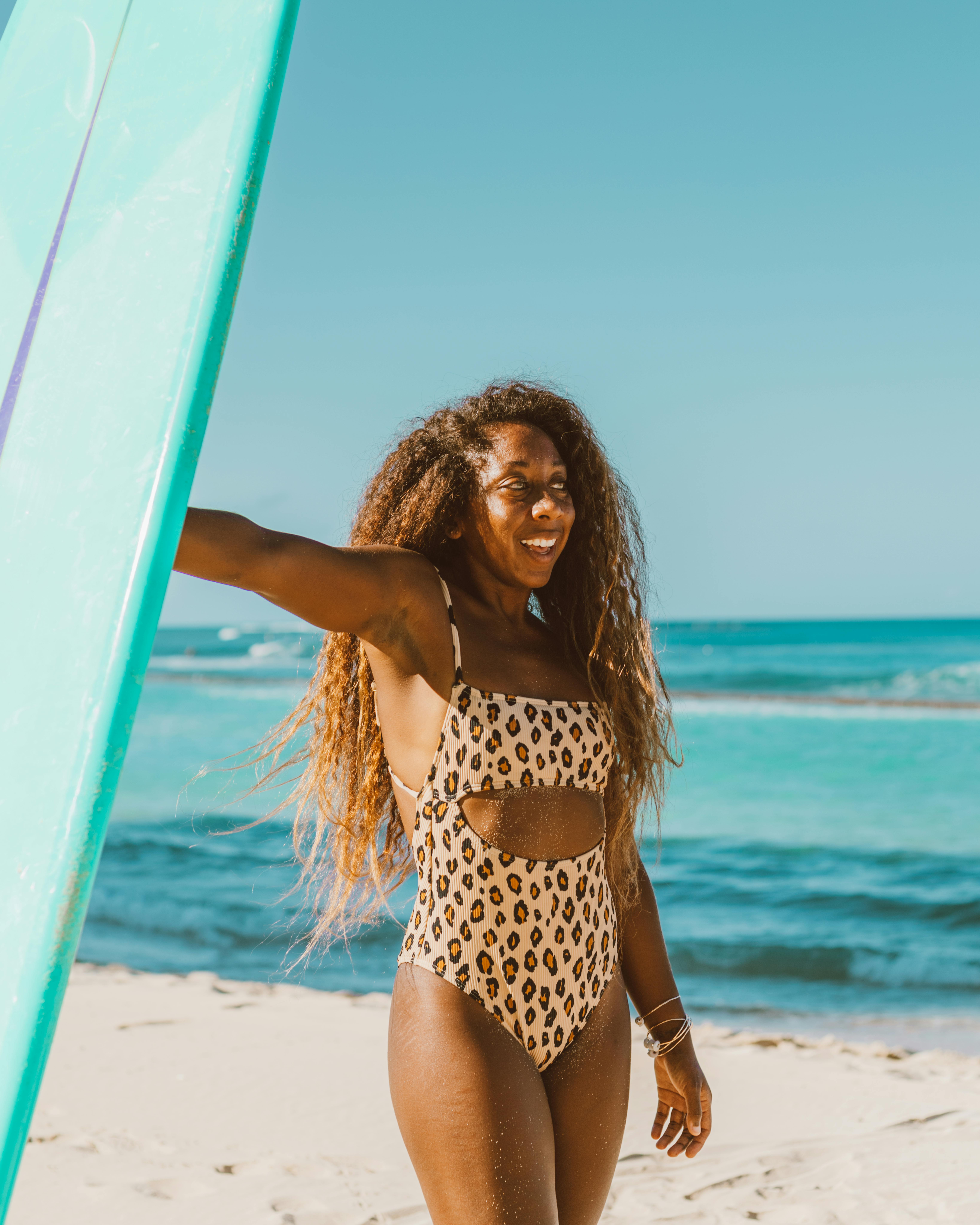 穿着豹纹泳衣的非裔美国妇女在阳光明媚的海滩上与冲浪板合影