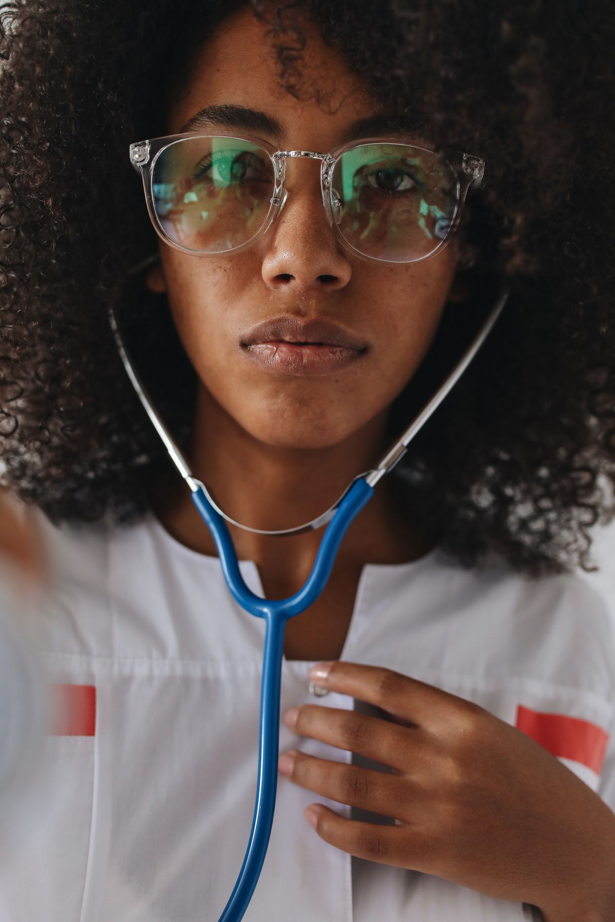 A Black woman doctor in a white coat and clear eyeglasses holds the chest piece of a blue stethoscope to her own chest, as if listening to a heartbeat.