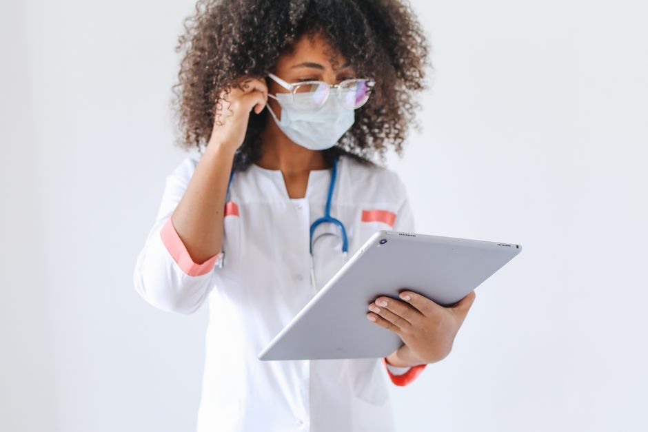 An African American female doctor wearing glasses and a face mask uses a tablet inside a medical facility.