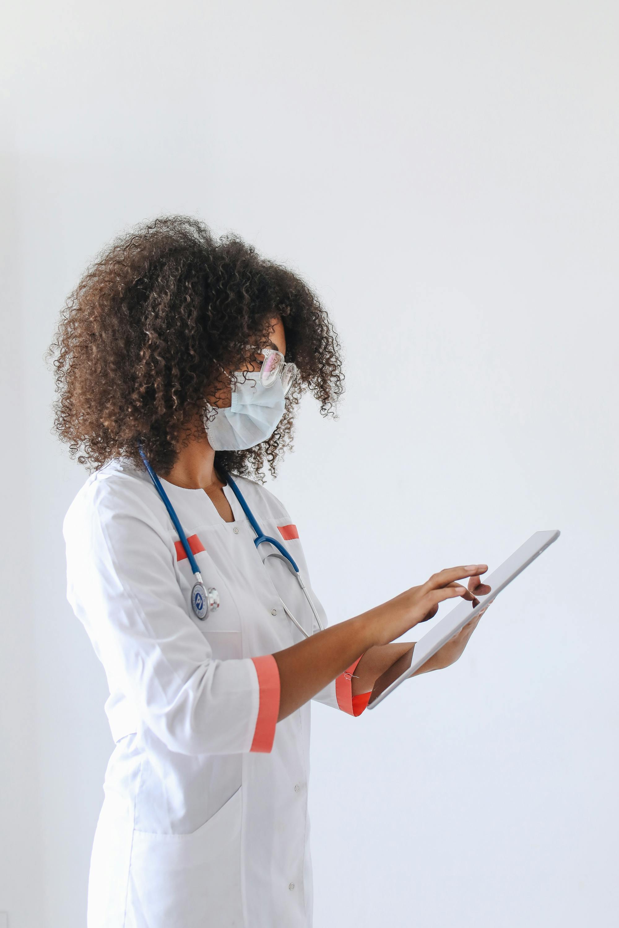 A female doctor in a white coat and face mask using a tablet for data entry in a medical setting.