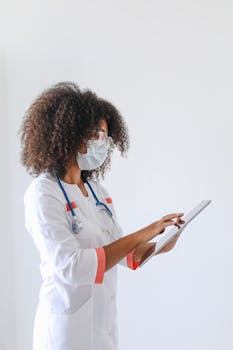 A female doctor in a white coat and face mask using a tablet for data entry in a medical setting.