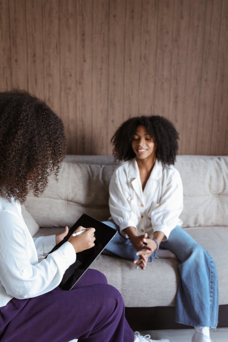 A Woman Sitting On The Couch Talking To The Woman Holding A Tablet