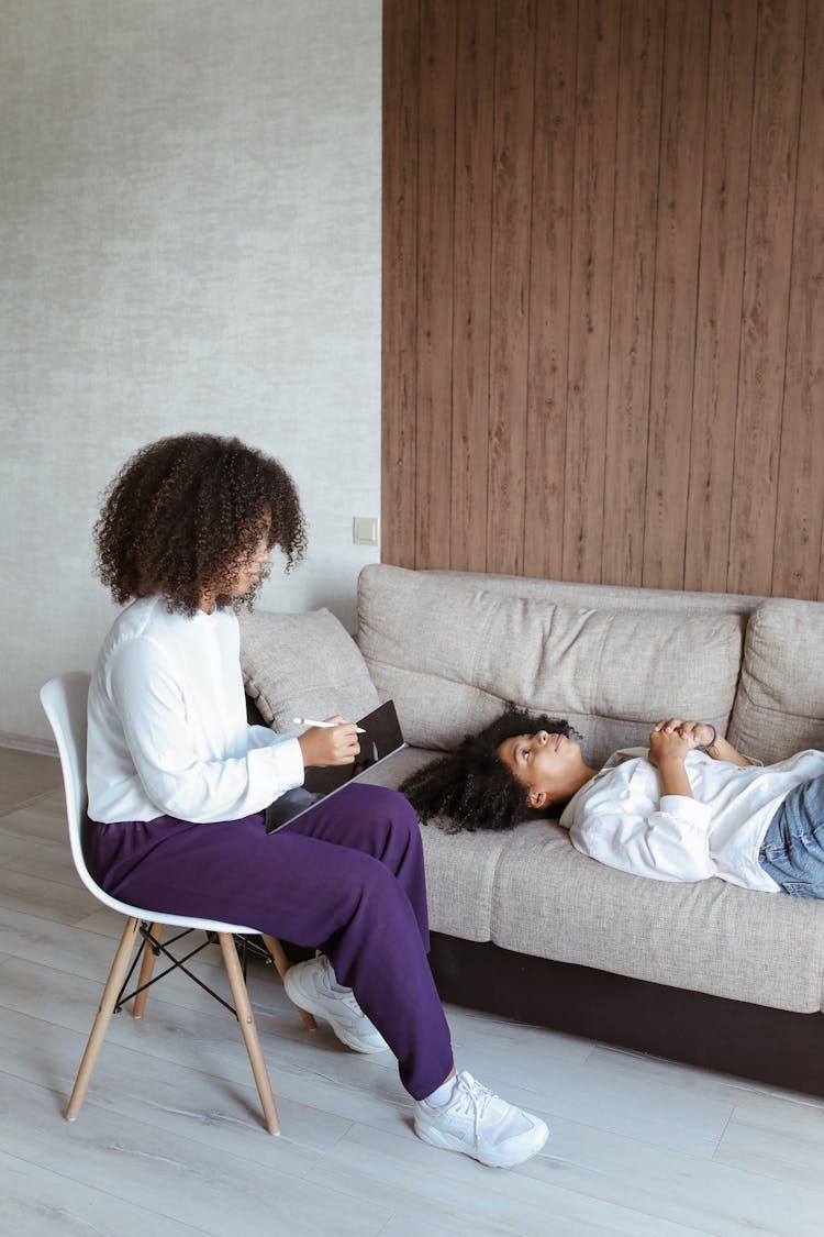 A Woman In White Long Sleeves Holding A Tablet While Talking To The Woman Lying On The Couch