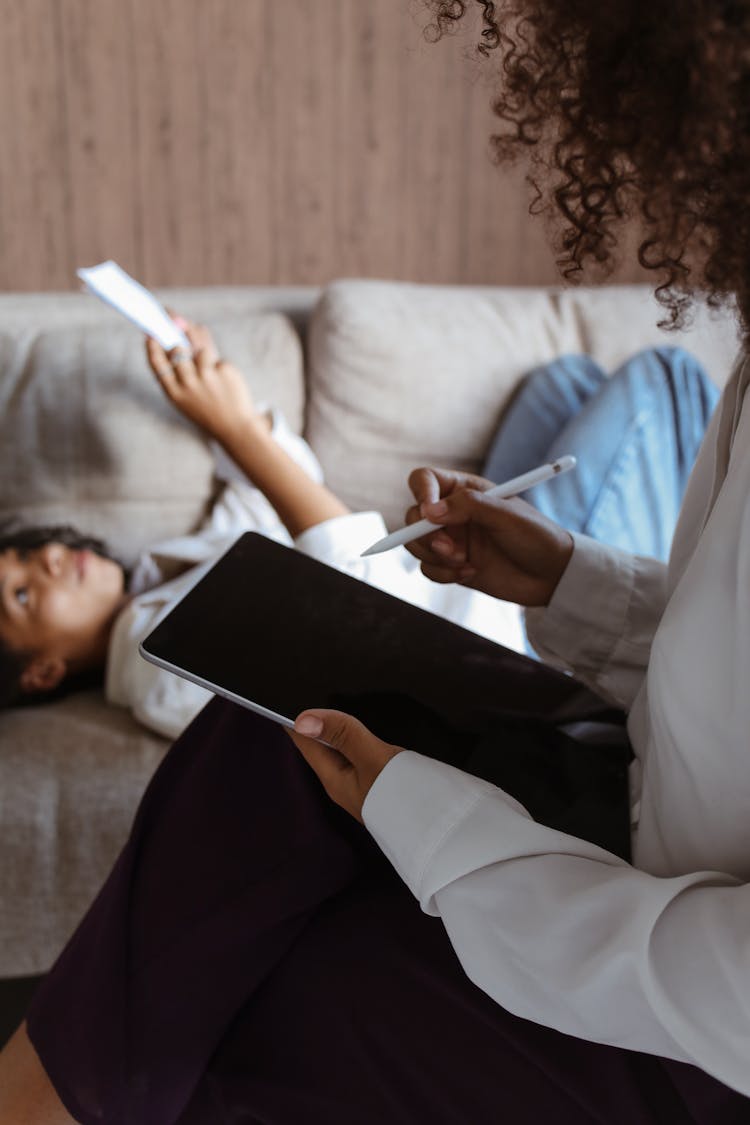 Woman Writing On A Tablet Device Using Stylus Pen Beside A Girl Lying On A Couch While Reading A Document