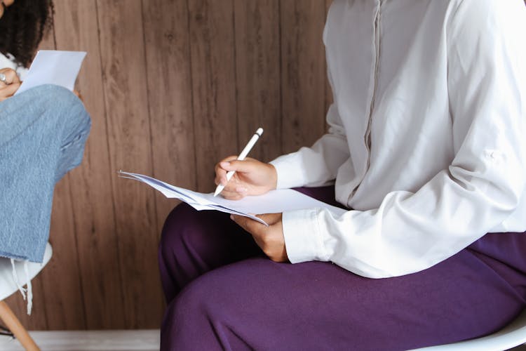 A Woman In White Long Sleeves Sitting On A Chair While Holding A Pen And A Document