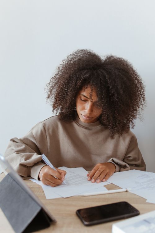 Free Young woman with curly hair writing notes in a home office setting with natural light. Stock Photo