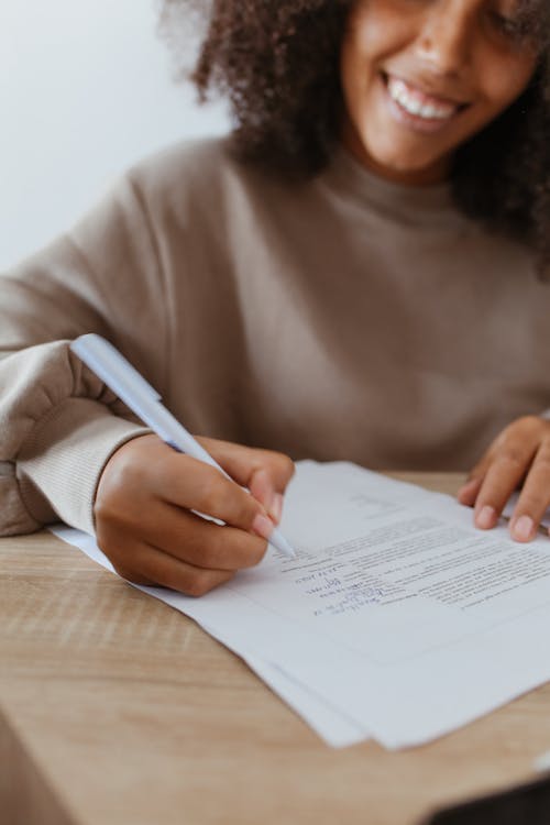 Woman in Brown Long Sleeve Shirt Holding a Pen and Writing on a Paper