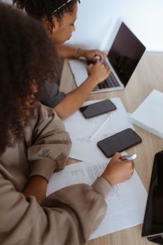Two young women collaborating at a workspace with laptops and smartphones indoors.