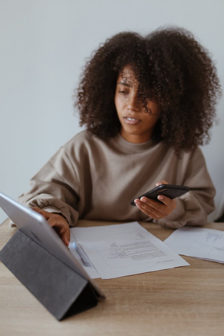 Woman In Brown Long Sleeve Shirt Holding Black Smartphone And Using A Tablet