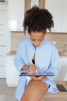 Young woman using a tablet with stylus in a bright modern kitchen, working remotely.