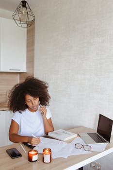 Young woman with curly hair working from home, surrounded by documents, candles, and a laptop.