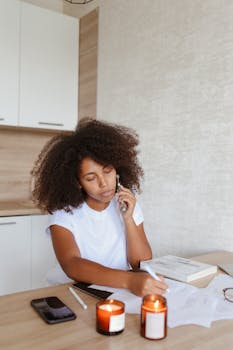 Professional woman multitasking at home, speaking on phone and writing, surrounded by cozy candles.