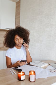 Adult woman with afro hair working remotely from home with smartphone, surrounded by candles and documents.