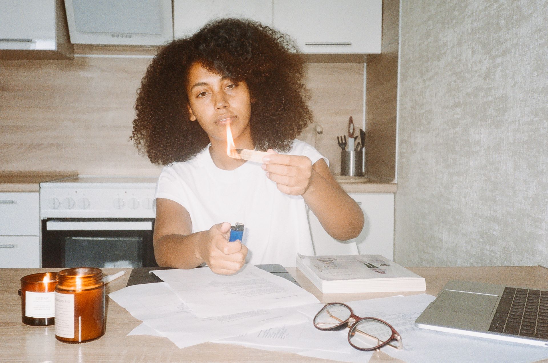 A woman with afro hair lights incense while working from home with a laptop on a kitchen counter.